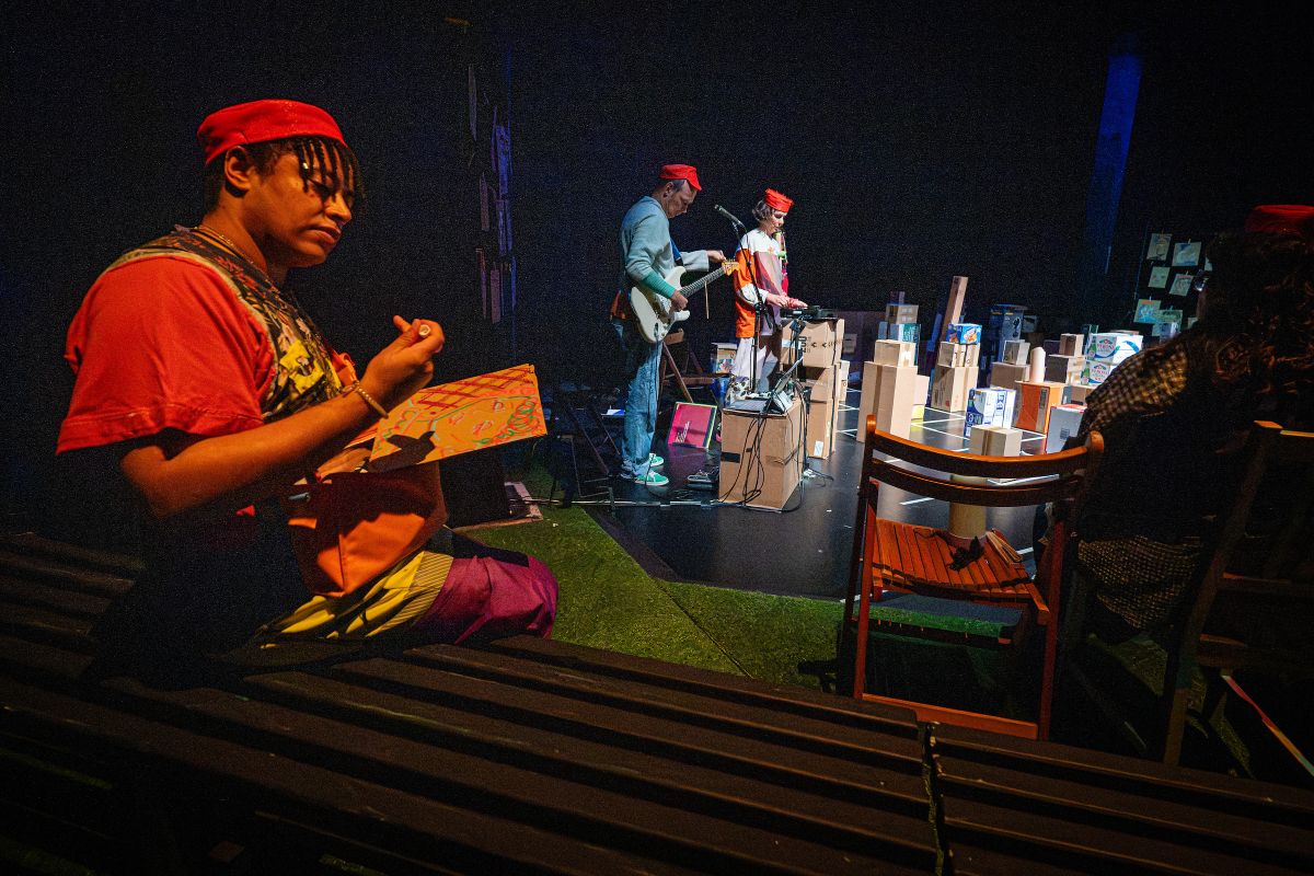 Person holding artwork sits beside benches as musicians perform among cardboard boxes on a theatre stage.