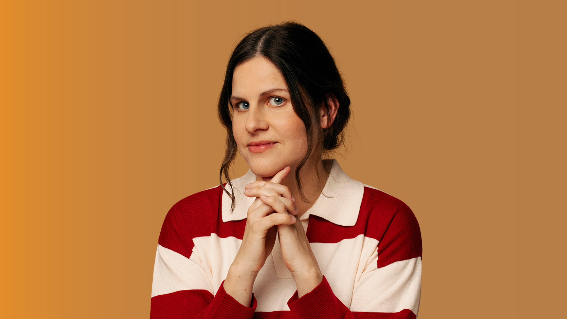 Portrait of a person with dark hair wearing a red and white striped collared jumper, posed with clasped hands against a plain warm brown background.