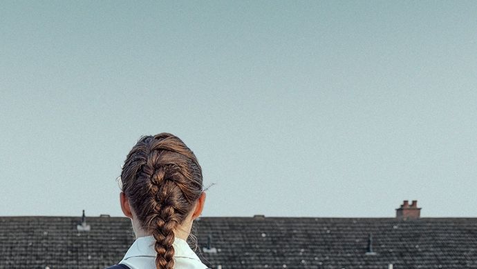 Person with a backpack stands facing a row of houses, with a book visible in the backpack pocket and open sky above.