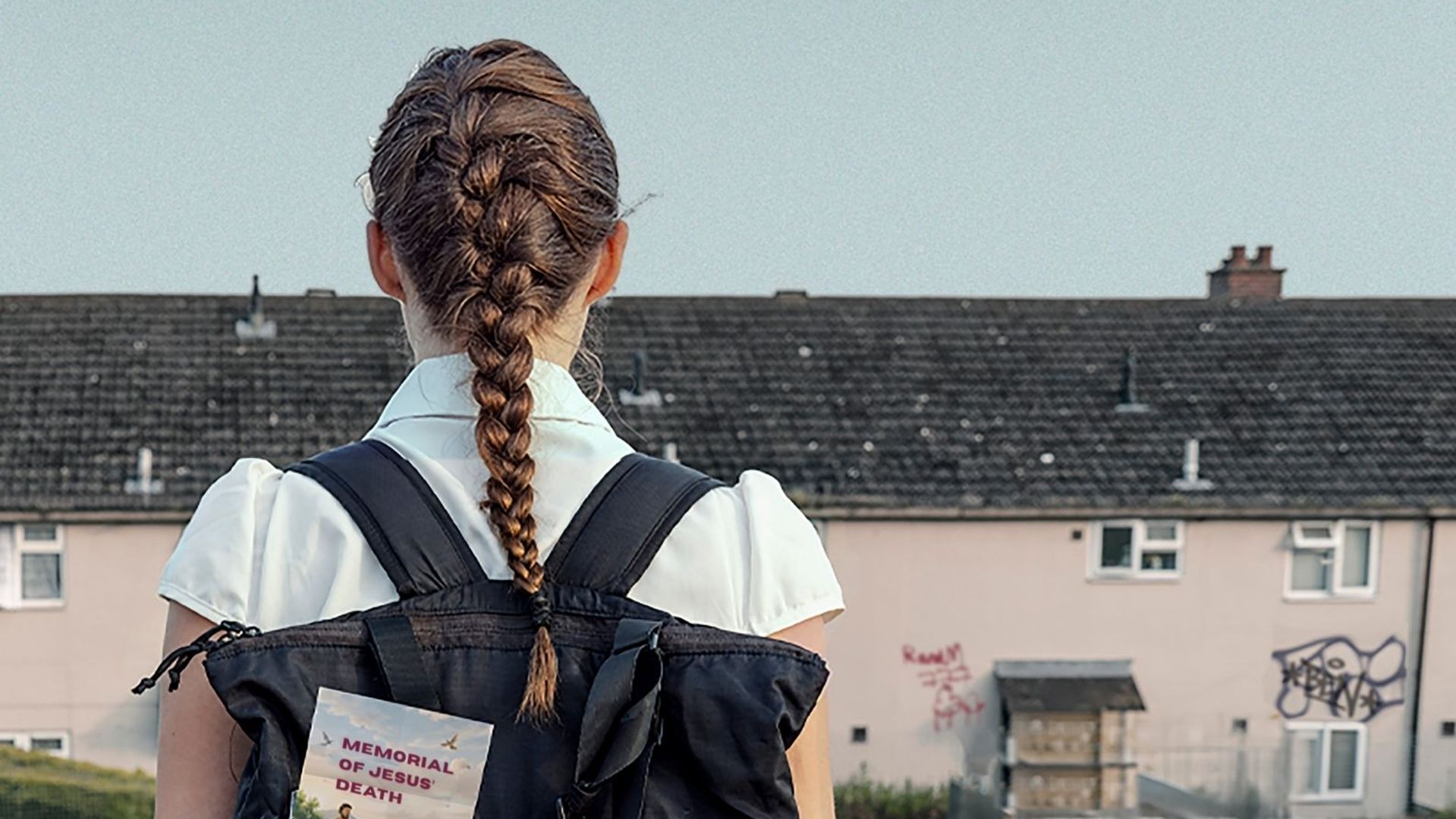 Person with a backpack stands facing a row of houses, with a book visible in the backpack pocket and open sky above.