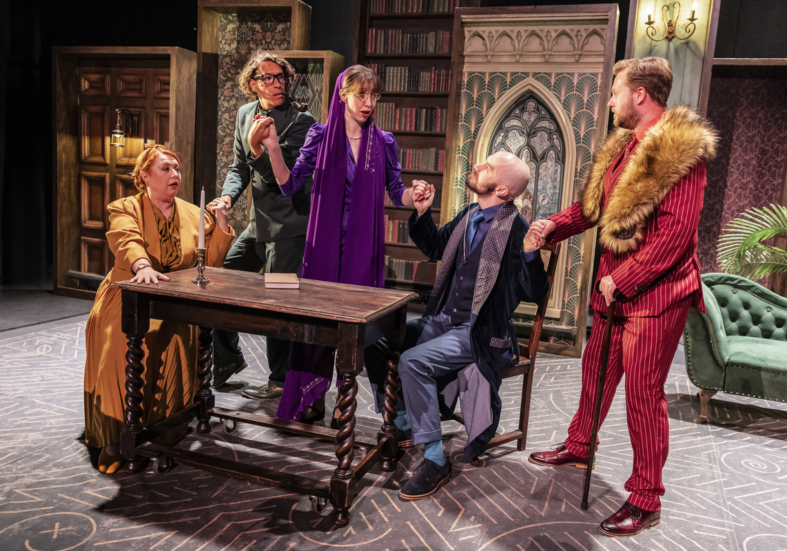 Five performers gather around a table in a mock séance scene, surrounded by bookshelves and vintage décor.