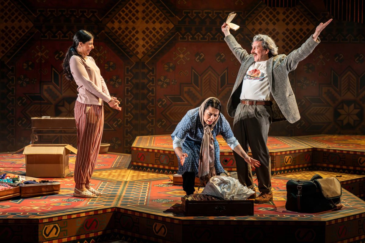 Three characters on stage expressing varied emotions. Laila’s father Hakim stands joyfully with raised arms, whilst Laila appears amused, and her mother crouches down over a suitcase, seemingly distressed.