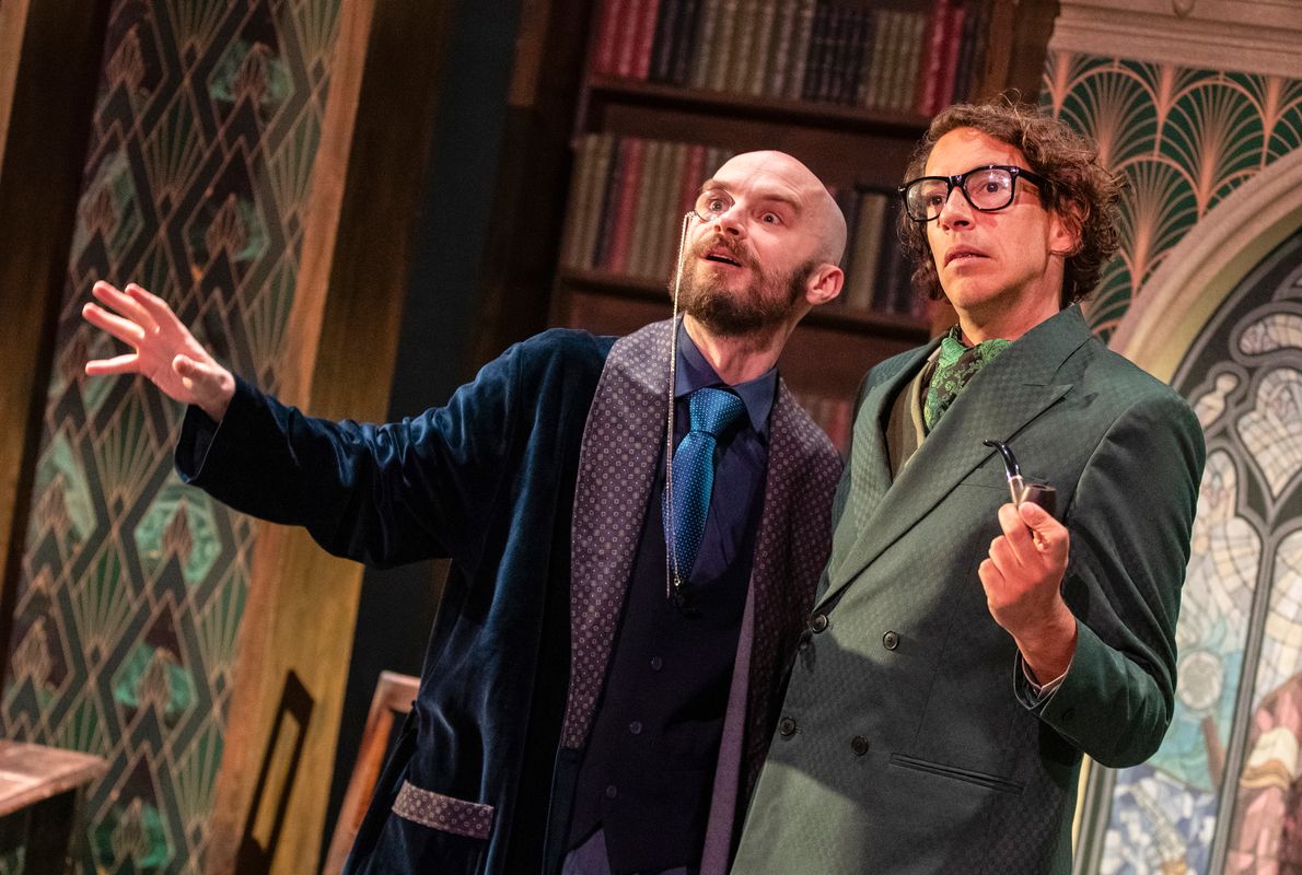 Two performers in theatrical detective-style costumes stand together in a library setting, one holding a pipe and the other gesturing ahead.