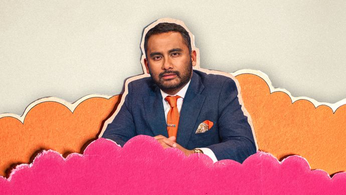 Person in a blue suit with an orange tie seated among layered pink and orange paper-cut clouds against a textured light background.