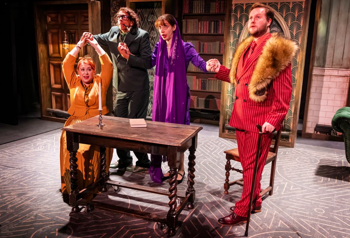 Four performers gather around a table in a séance-style scene, holding hands in a library setting with vintage décor.