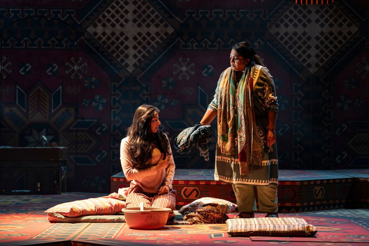 Mariam is handing a traditional Afghani dress to Laila, who is seated to the floor, in casual Westernised clothing.