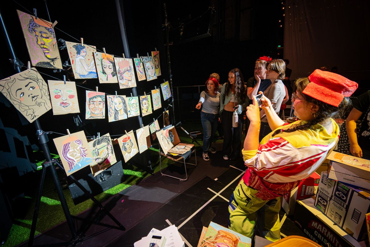 People view and photograph a display of illustrated portraits hanging on lines beside stacked cardboard boxes.