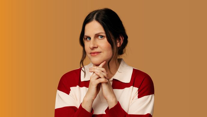Portrait of a person with dark hair wearing a red and white striped collared jumper, posed with clasped hands against a plain warm brown background.