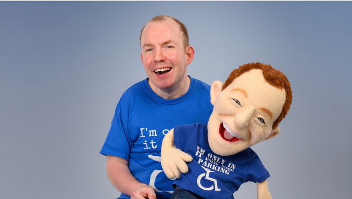 Performer sits beside a large puppet, both wearing matching blue T-shirts against grey backdrop.