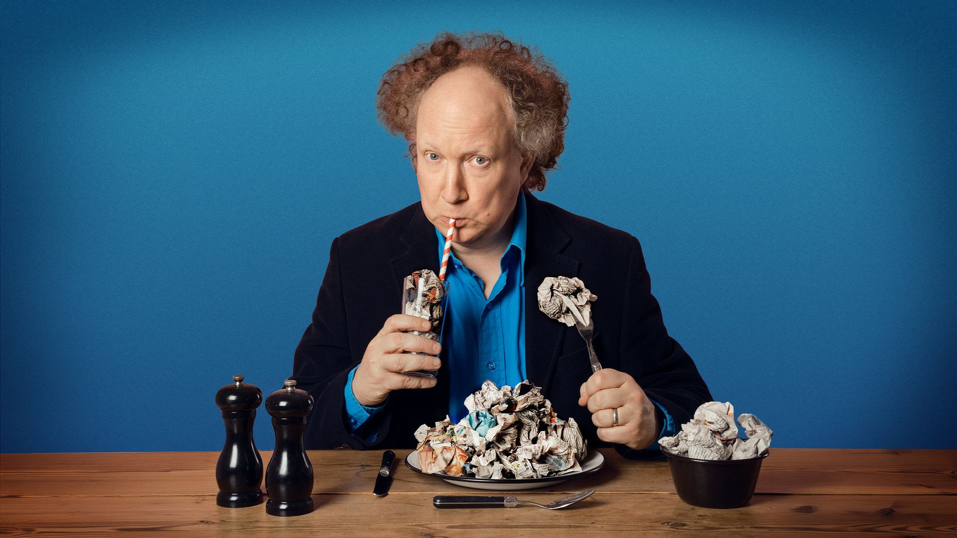 Performer sits at a table drinking from a carton, surrounded by crumpled paper balls.