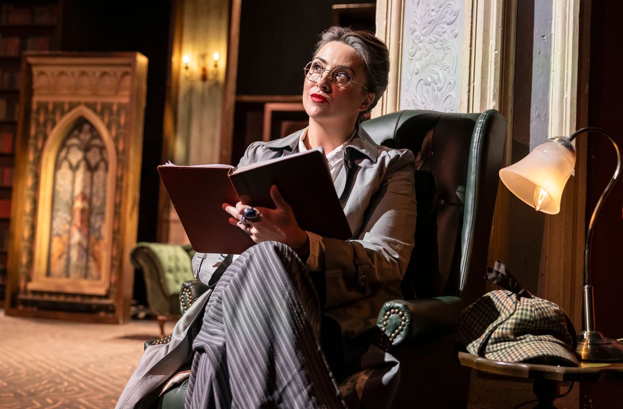 Performer seated in a leather armchair with an open notebook, surrounded by bookshelves and stained-glass-style panels.