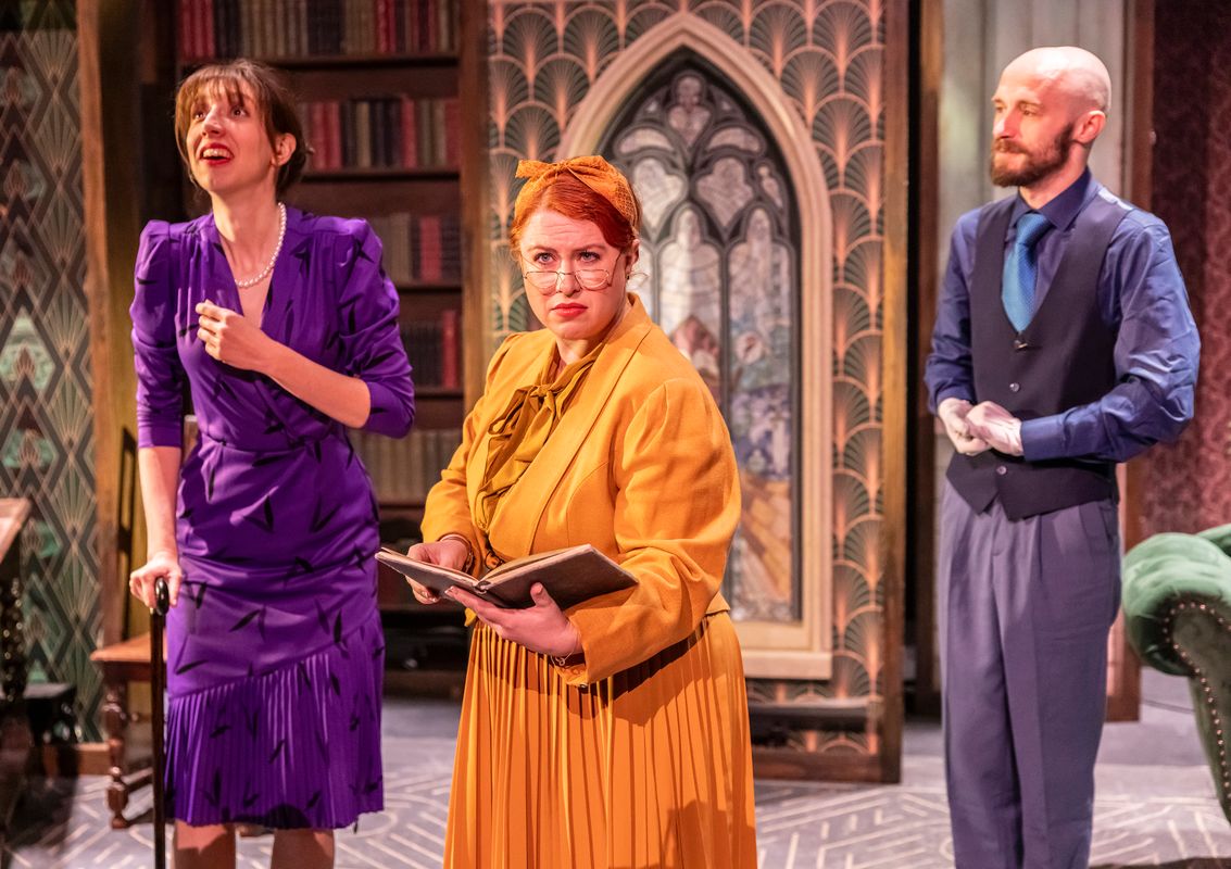 Three performers in colourful period-style costumes examine a notebook in a library setting with stained-glass-style panels behind them.