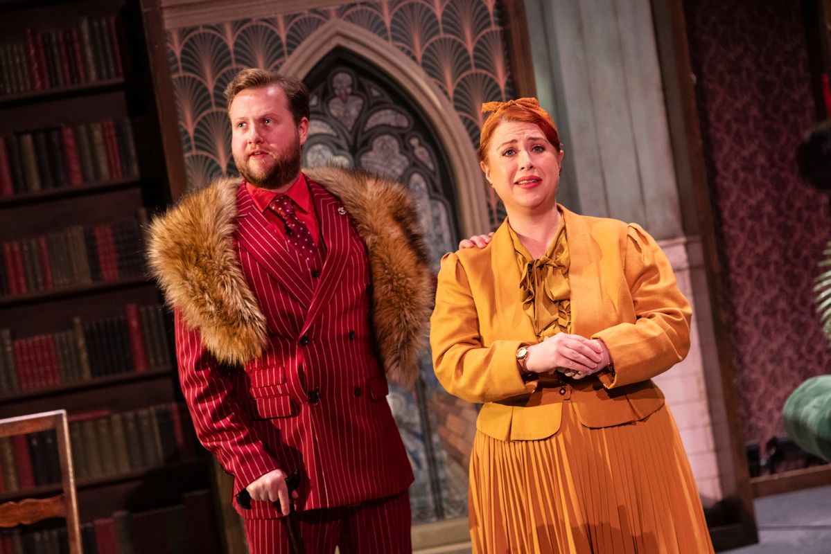 Two performers in colourful period-style costumes stand in a library setting, with bookshelves and stained-glass-style panels behind them.